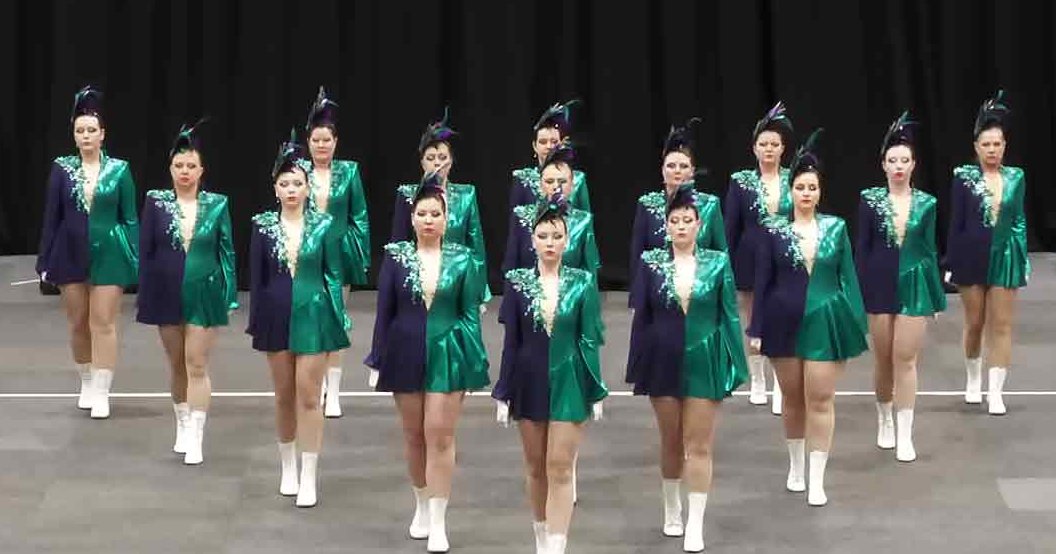 Young adult female dancers performing in a studio setting, wearing shiny green jackets with white accents and matching skirts, white ankle boots, and feathered headpieces.