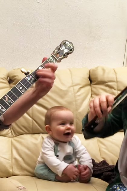 A family scene indoors with an adult male, middle-aged infant, and adult female playing musical instruments on a couch under soft lighting.