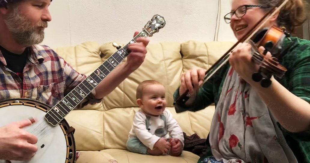 A family scene indoors with an adult male, middle-aged infant, and adult female playing musical instruments on a couch under soft lighting.