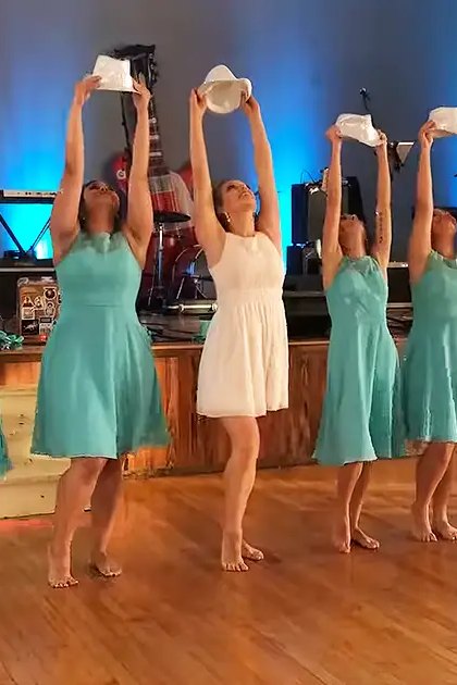 A dance group of young adult females performing on a wooden floor with blue stage lighting, likely an indoor hall or studio at night.
