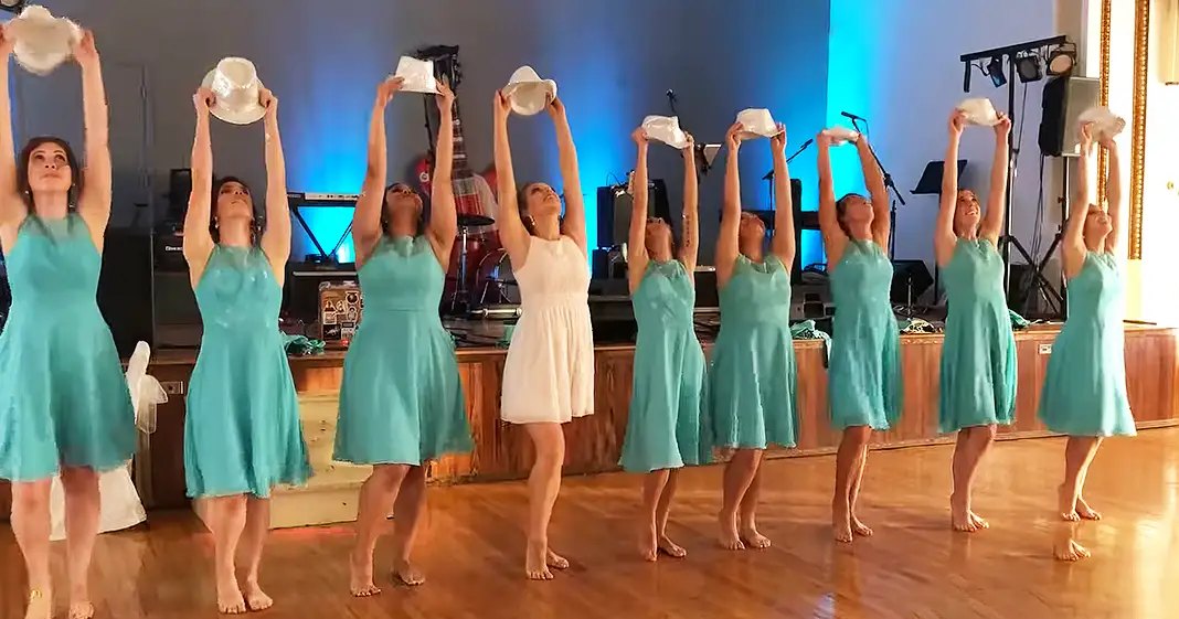 A dance group of young adult females performing on a wooden floor with blue stage lighting, likely an indoor hall or studio at night.