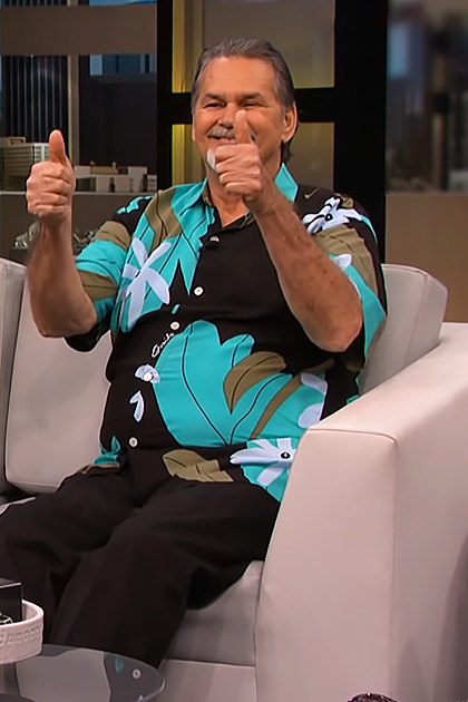 Two older adult males seated on a white couch in a modern studio with dark panels and soft lighting, wearing patterned shirts and beige pants.
