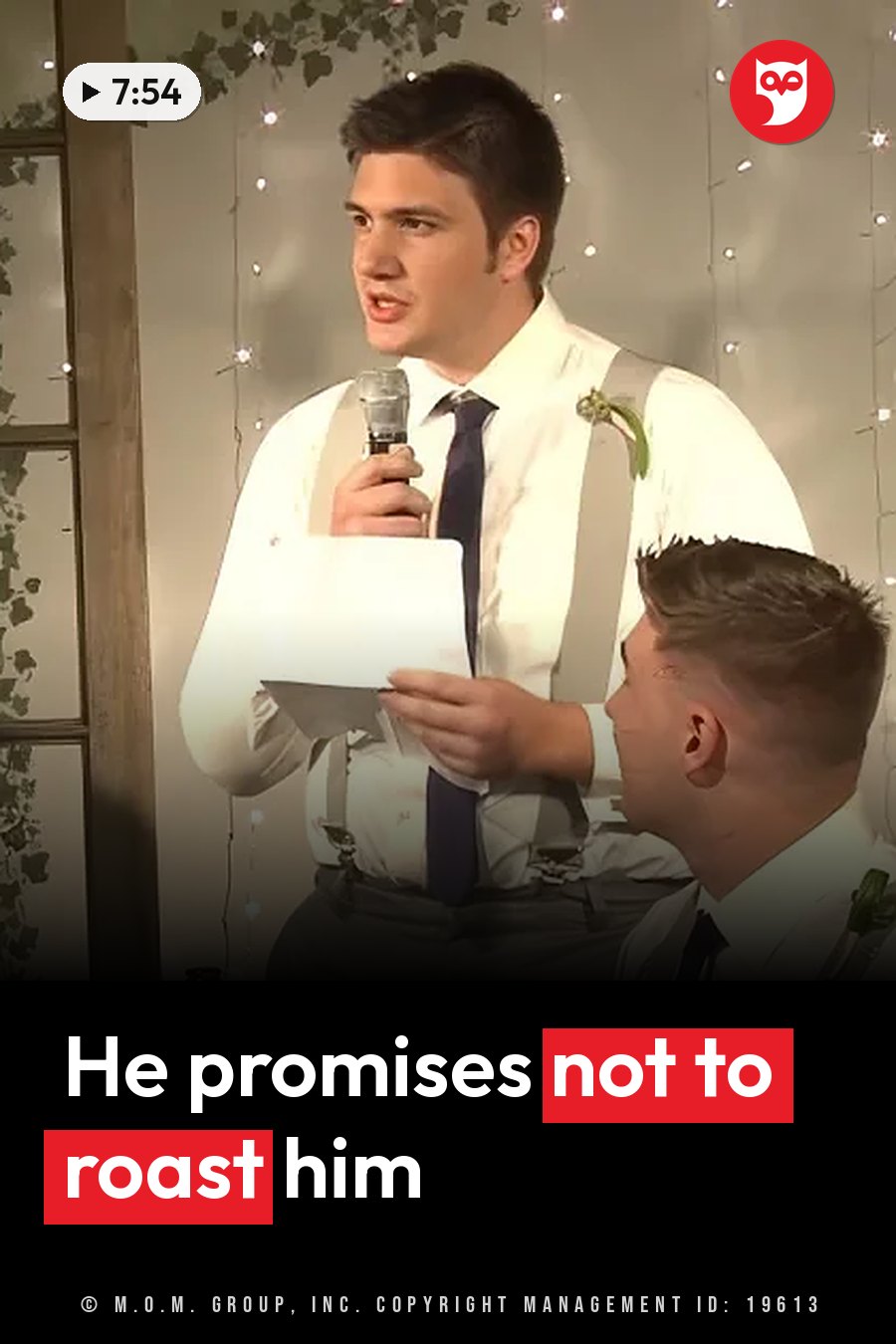 A man in a white shirt and tie holds a microphone and a sheet of paper as he speaks, with string lights behind him. A headline runs in white letters across the lower third.