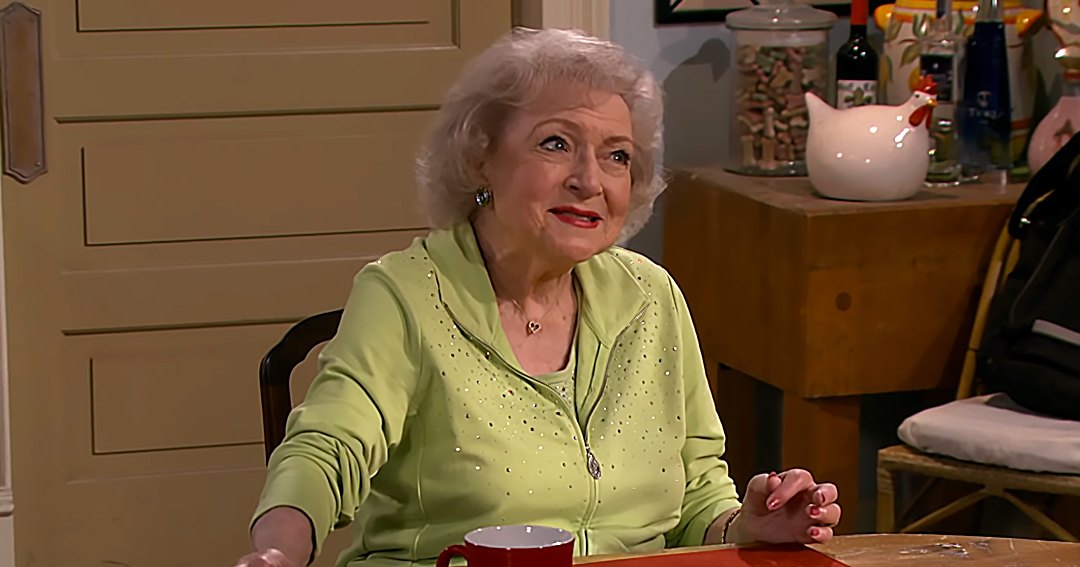 An older adult female wearing a light green zip-up cardigan with small sequins and a heart pendant necklace, sitting at a wooden table in a cozy kitchen setting with red placemat and various home decor items.