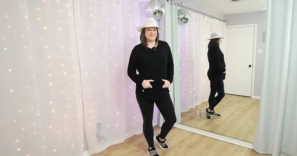 A young adult female in a casual outfit stands indoors, with white sheer curtains and a disco ball reflecting light from the ceiling.