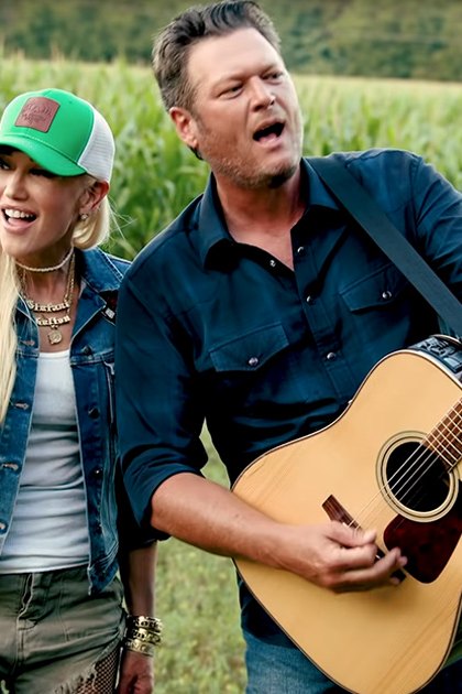 A musical duo in an outdoor rural setting with cornfields, late summer or early fall attire, soft morning or evening light. Both individuals wear bracelets and layered necklaces; one holds a light brown acoustic guitar.