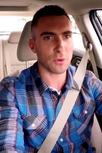 An adult male wearing a blue plaid button-down shirt with cotton fabric and accessories, seated in the driver's seat of a modern car with beige leather seats and quilted patterns. The scene takes place inside an urban area during daytime.