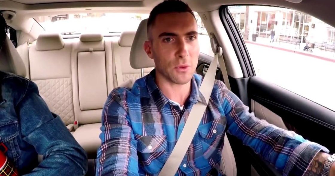 An adult male wearing a blue plaid button-down shirt with cotton fabric and accessories, seated in the driver's seat of a modern car with beige leather seats and quilted patterns. The scene takes place inside an urban area during daytime.