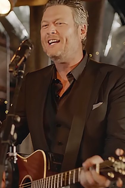 A middle-aged male singer in a dark suit jacket and black shirt plays guitar on stage with string lights and rustic wooden paneling, accompanied by a young adult female performer in an off-the-shoulder dress. Warm lighting creates an inviting ambiance.