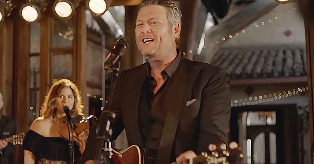 A middle-aged male singer in a dark suit jacket and black shirt plays guitar on stage with string lights and rustic wooden paneling, accompanied by a young adult female performer in an off-the-shoulder dress. Warm lighting creates an inviting ambiance.
