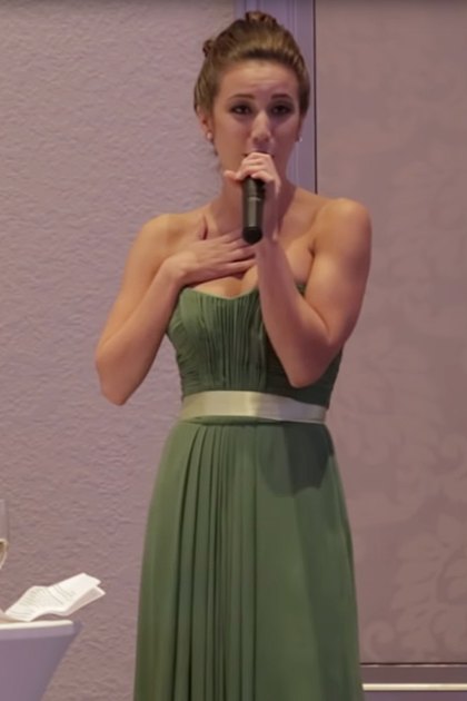 A young adult female in a green sleeveless floor-length gown with a gold belt, giving a speech at an indoor wedding reception alongside a smiling bride and seated groom in a gray suit during the evening.
