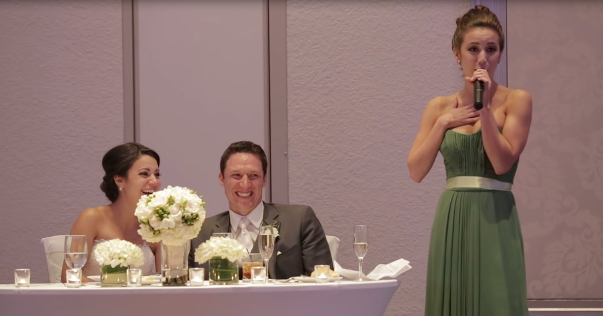 A young adult female in a green sleeveless floor-length gown with a gold belt, giving a speech at an indoor wedding reception alongside a smiling bride and seated groom in a gray suit during the evening.