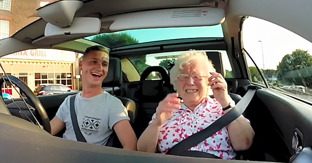 Young adult male driver wearing a light gray t-shirt with an Aztec-inspired pattern, accompanied by an older female passenger in a white blouse with pink floral patterns inside a car parked near 'China Grill' restaurant during the daytime.