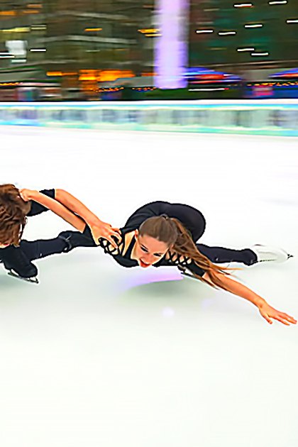 Young adult male and female figure skaters in a well-lit indoor ice rink with smooth skating surface, wearing fitted black attire and white skate boots.