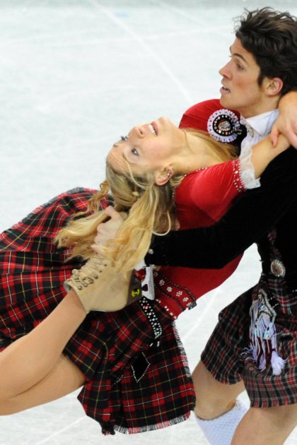 A young female figure skater in a red and black plaid skirt and white lace top performs alongside an adult male partner at an indoor ice rink, both dressed in figure skating attire.