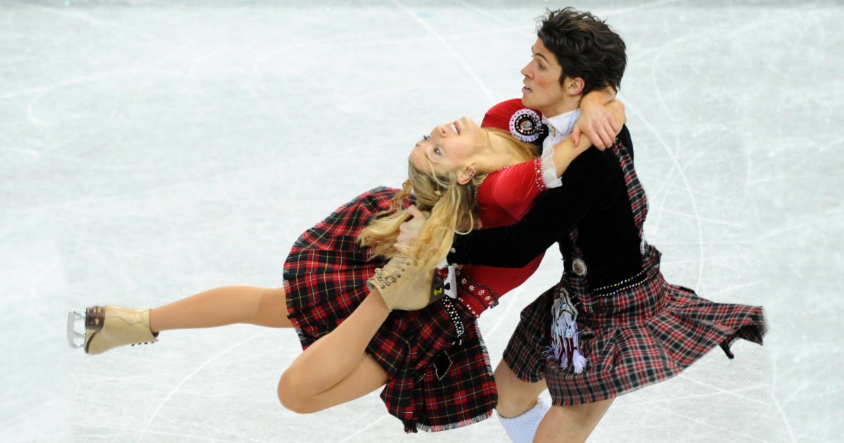 A young female figure skater in a red and black plaid skirt and white lace top performs alongside an adult male partner at an indoor ice rink, both dressed in figure skating attire.