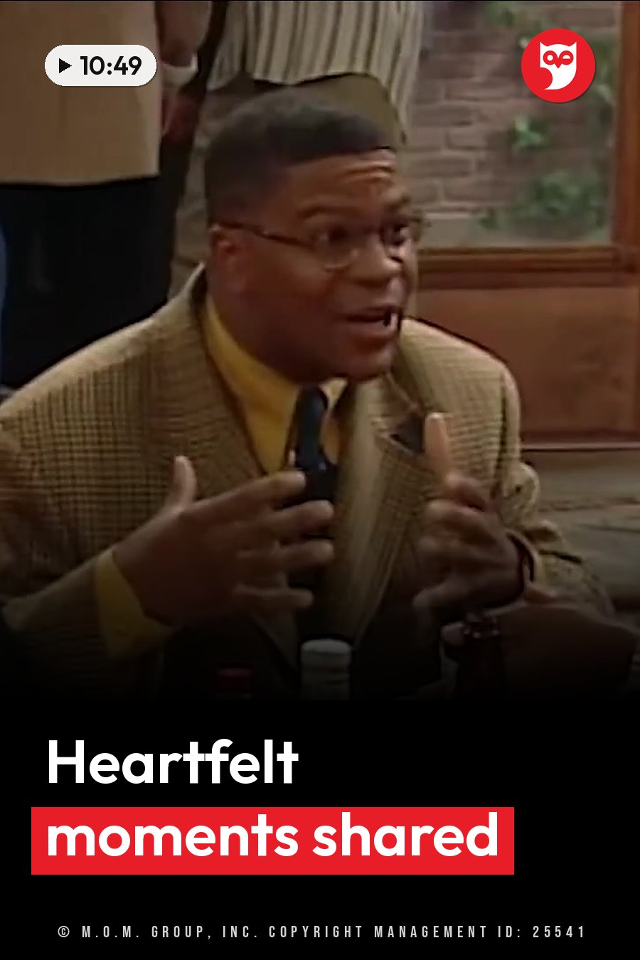 A man in glasses, a yellow shirt and a patterned jacket gestures with his hands while speaking into a microphone. A headline runs in white letters across the lower third.