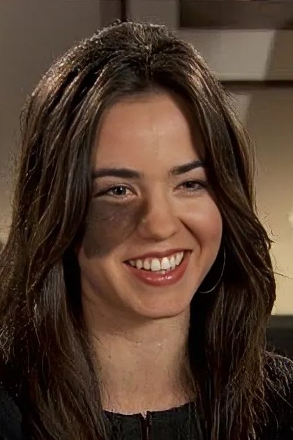 A young adult female wearing a dark-colored top with short sleeves in an indoor setting with furniture like a table with a lamp and vase of white flowers, suggesting a comfortable living space.