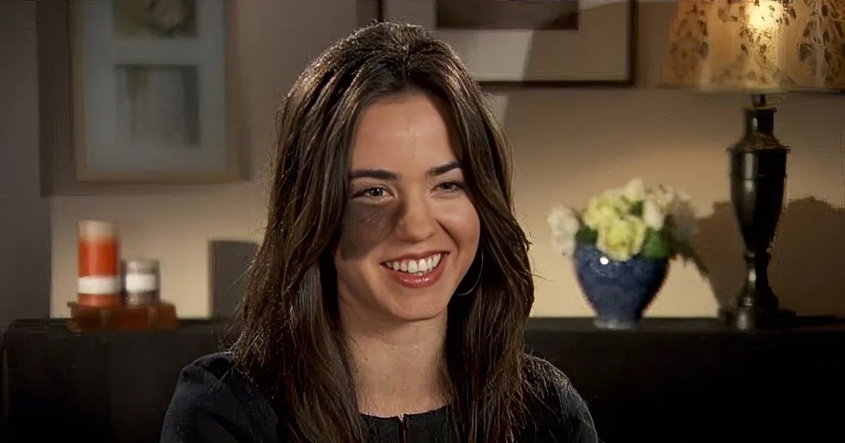 A young adult female wearing a dark-colored top with short sleeves in an indoor setting with furniture like a table with a lamp and vase of white flowers, suggesting a comfortable living space.