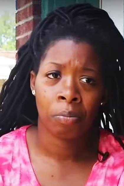 A middle-aged adult female wears a relaxed fit, pink and white tie-dye V-neck short-sleeved shirt with small earrings on an urban brick wall background.