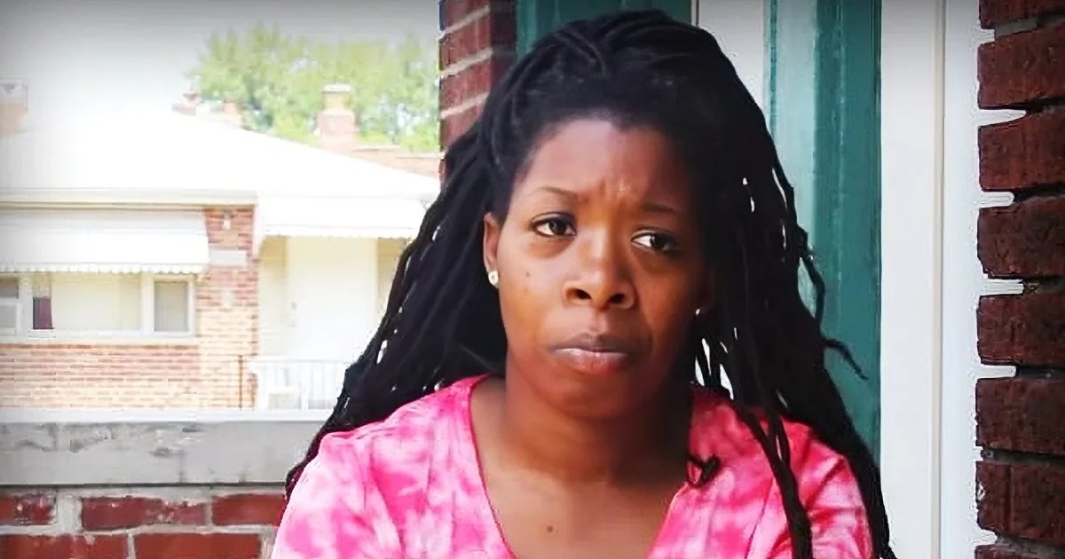 A middle-aged adult female wears a relaxed fit, pink and white tie-dye V-neck short-sleeved shirt with small earrings on an urban brick wall background.