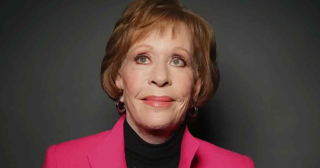 An older adult female wearing a bright pink blazer with notched lapels and black turtleneck, holding 'Madly Odd' text in a studio setting.