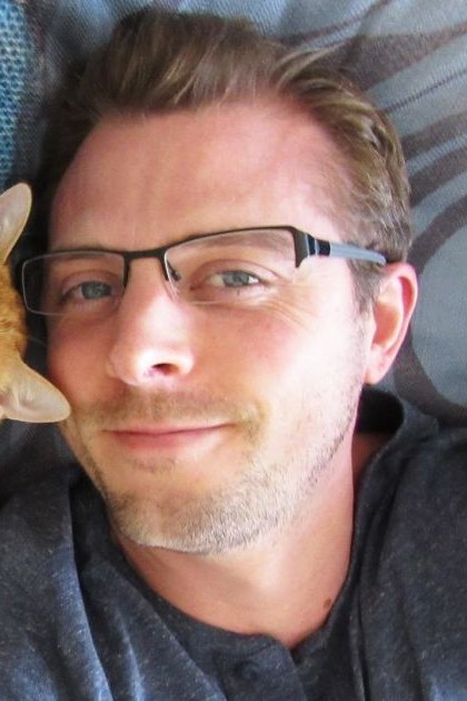 An adult male wearing a dark gray t-shirt, lying with an orange tabby cat on blue patterned fabric in a home setting during daytime.