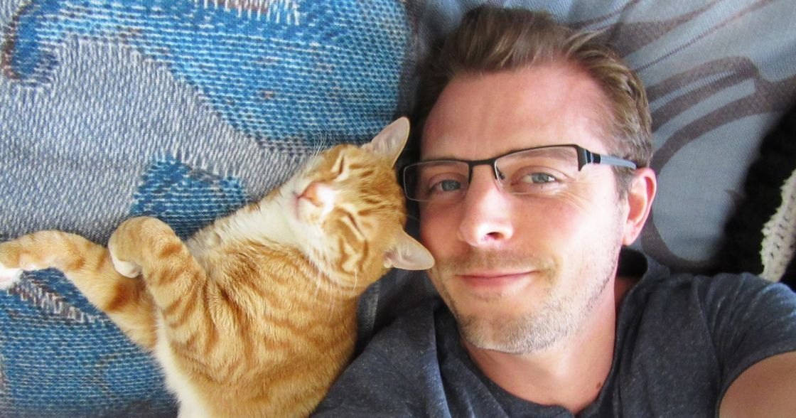 An adult male wearing a dark gray t-shirt, lying with an orange tabby cat on blue patterned fabric in a home setting during daytime.
