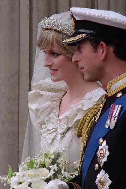 A young adult female wearing a white bridal gown with intricate lace detailing, standing alongside an adult male in a black military uniform at what appears to be Westminster Abbey during daylight.