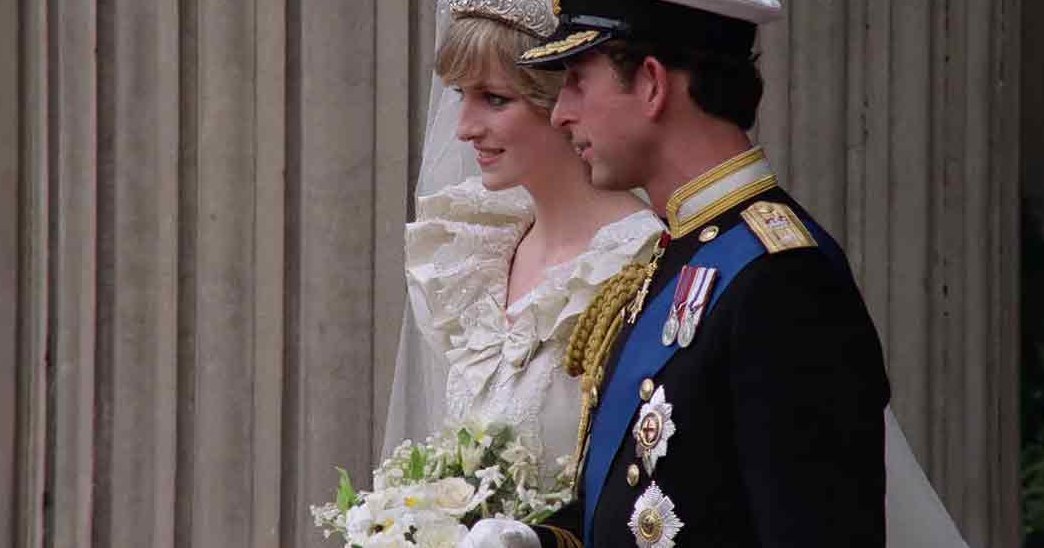 A young adult female wearing a white bridal gown with intricate lace detailing, standing alongside an adult male in a black military uniform at what appears to be Westminster Abbey during daylight.