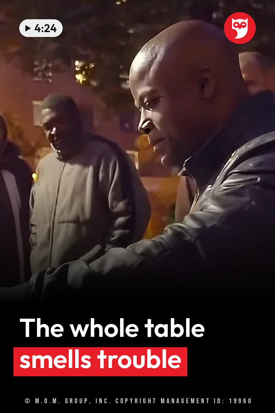 A bald man in a leather jacket looks down while other people stand nearby outdoors at night. A headline runs in white letters across the lower third.