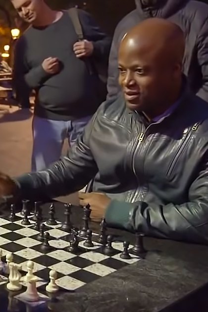 Adults playing chess in a park at night, surrounded by benches, trees, and artificial lighting.