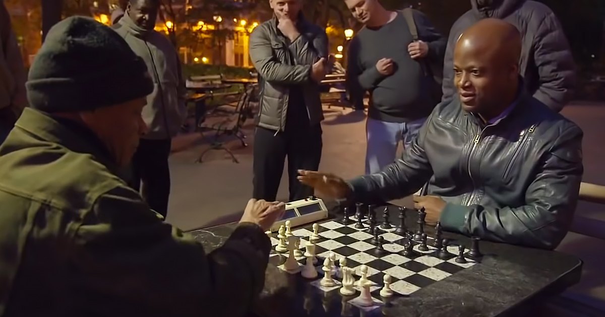 Adults playing chess in a park at night, surrounded by benches, trees, and artificial lighting.