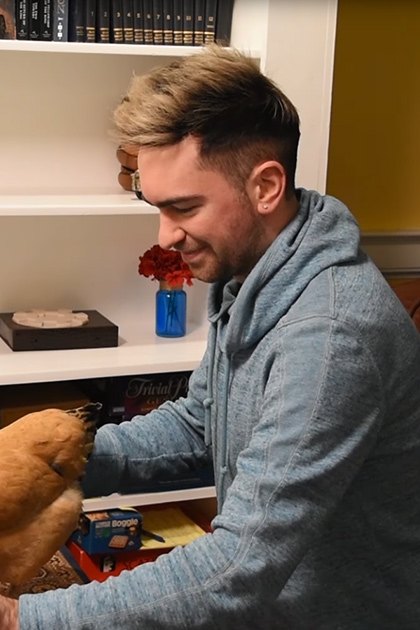 A Rhode Island Red chicken perched on a keyboard being played by an adult male in a home office or living room setting with white bookshelf, dark leather couch, and yellow walls.