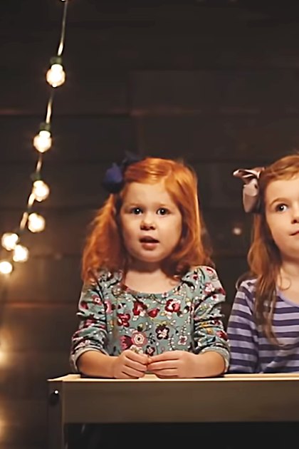 Two young female children in a cozy indoor setting, wearing floral and striped dresses under string lights.