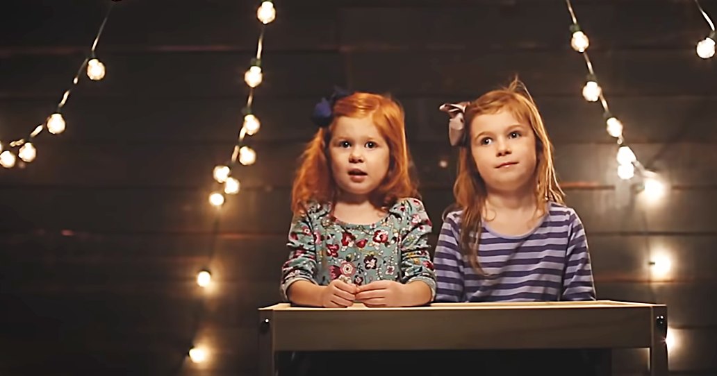 Two young female children in a cozy indoor setting, wearing floral and striped dresses under string lights.