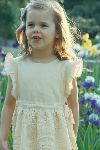 A toddler girl wearing a cream-colored lace dress with ruffled sleeves stands amidst blooming irises in an outdoor garden setting, surrounded by greenery and soft natural daylight.