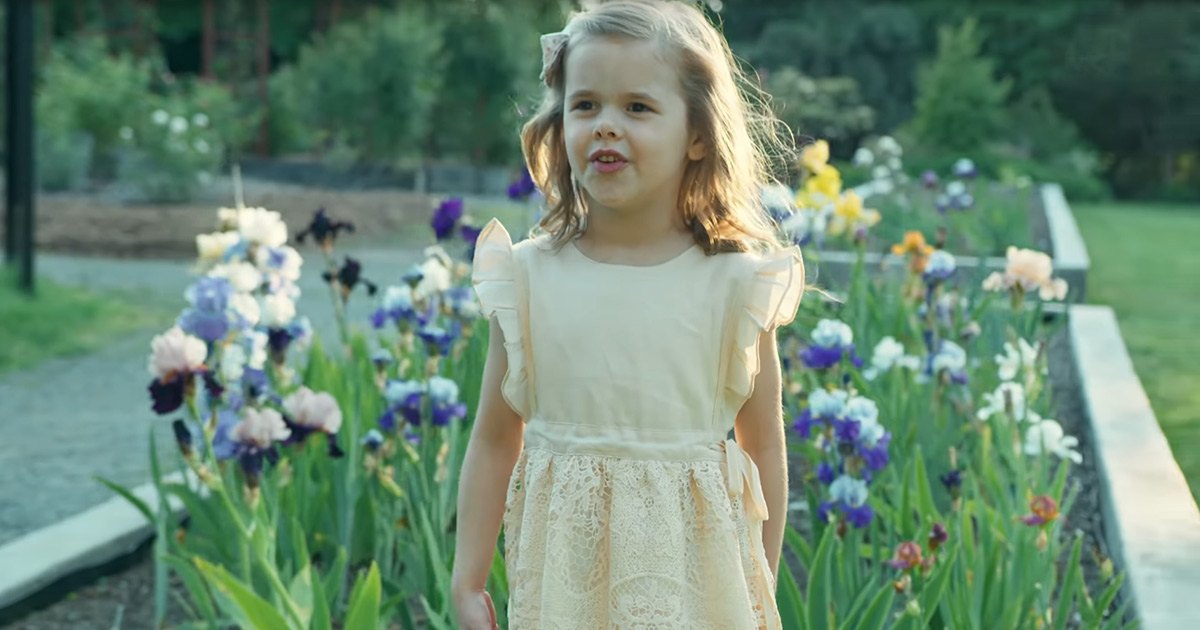 A toddler girl wearing a cream-colored lace dress with ruffled sleeves stands amidst blooming irises in an outdoor garden setting, surrounded by greenery and soft natural daylight.