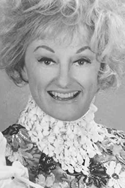 An older adult woman wearing a floral-patterned garment with intricate detailing and high collar or ruffled neckpiece in a studio setting, holding a cigarette.