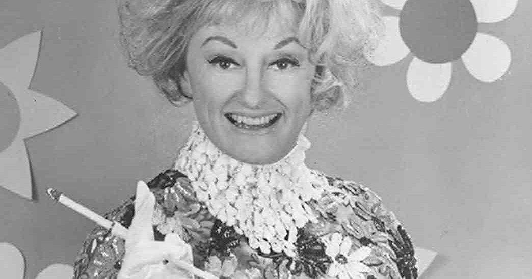 An older adult woman wearing a floral-patterned garment with intricate detailing and high collar or ruffled neckpiece in a studio setting, holding a cigarette.