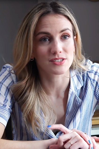 A young adult female in a relaxed, short-sleeved striped shirt with blue and white horizontal stripes, wearing small hoop earrings and rings, stands indoors by a wooden chair. The setting is a home environment with natural daylight.