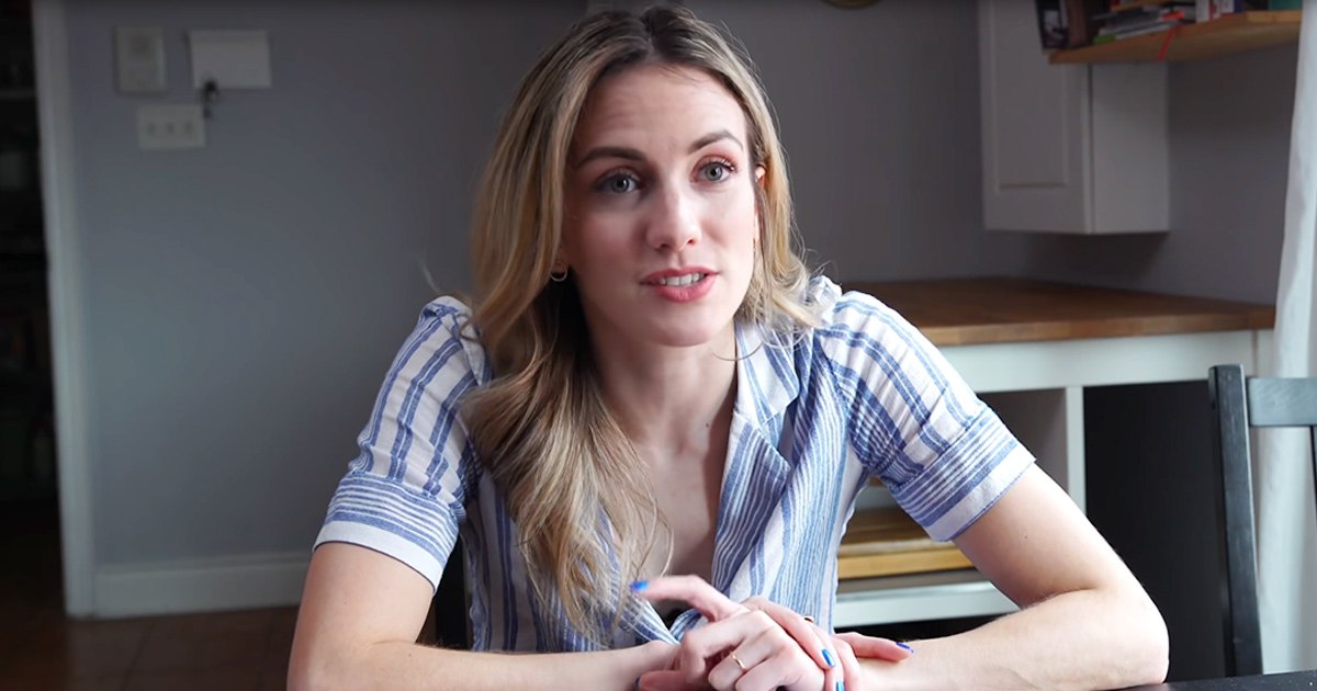 A young adult female in a relaxed, short-sleeved striped shirt with blue and white horizontal stripes, wearing small hoop earrings and rings, stands indoors by a wooden chair. The setting is a home environment with natural daylight.