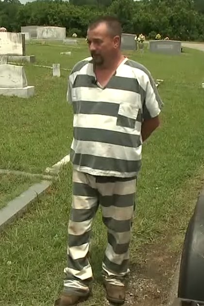 A middle-aged man wearing a prison jumpsuit with vertical gray and white stripes, standing in front of tombstones at a cemetery. The setting includes flowers on the tombstones, green grass, and daylight illuminating the scene.