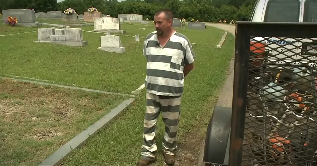 A middle-aged man wearing a prison jumpsuit with vertical gray and white stripes, standing in front of tombstones at a cemetery. The setting includes flowers on the tombstones, green grass, and daylight illuminating the scene.