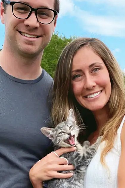 A young adult male singer and female holding a tabby kitten outdoors under clear blue skies; white car partially visible in the background, urban or suburban environment.