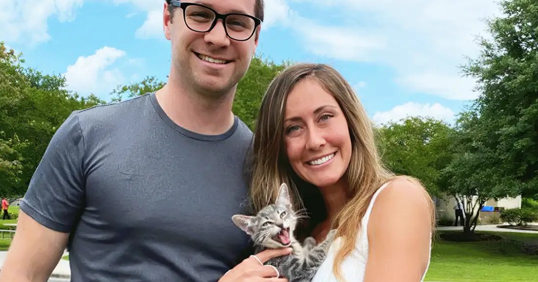 A young adult male singer and female holding a tabby kitten outdoors under clear blue skies; white car partially visible in the background, urban or suburban environment.
