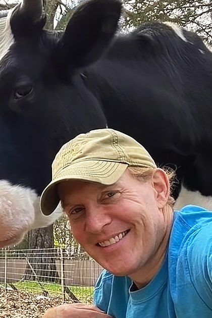 A young adult female with long reddish-brown hair takes a selfie in an outdoor rural setting with two cows, one brown and white, the other black-and-white Holstein. Trees and natural light suggest spring or summer.