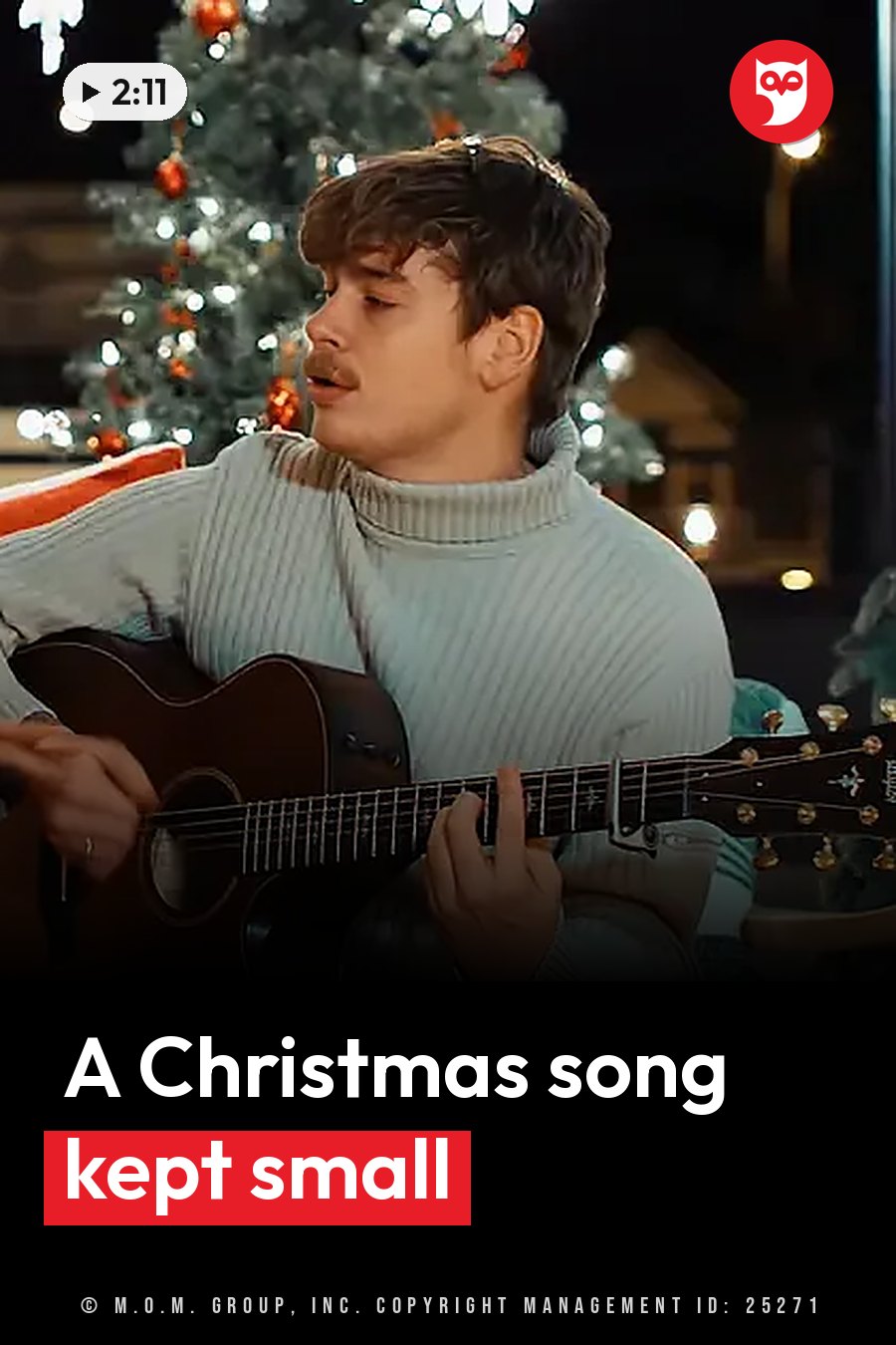 A man plays an acoustic guitar while singing, wearing a turtleneck sweater, with a lit Christmas tree behind him. A headline runs in white letters across the lower third.