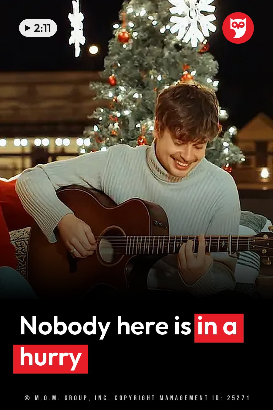 A man smiles as he plays guitar, seated in front of a decorated Christmas tree with lights at night. A headline runs in white letters across the lower third.