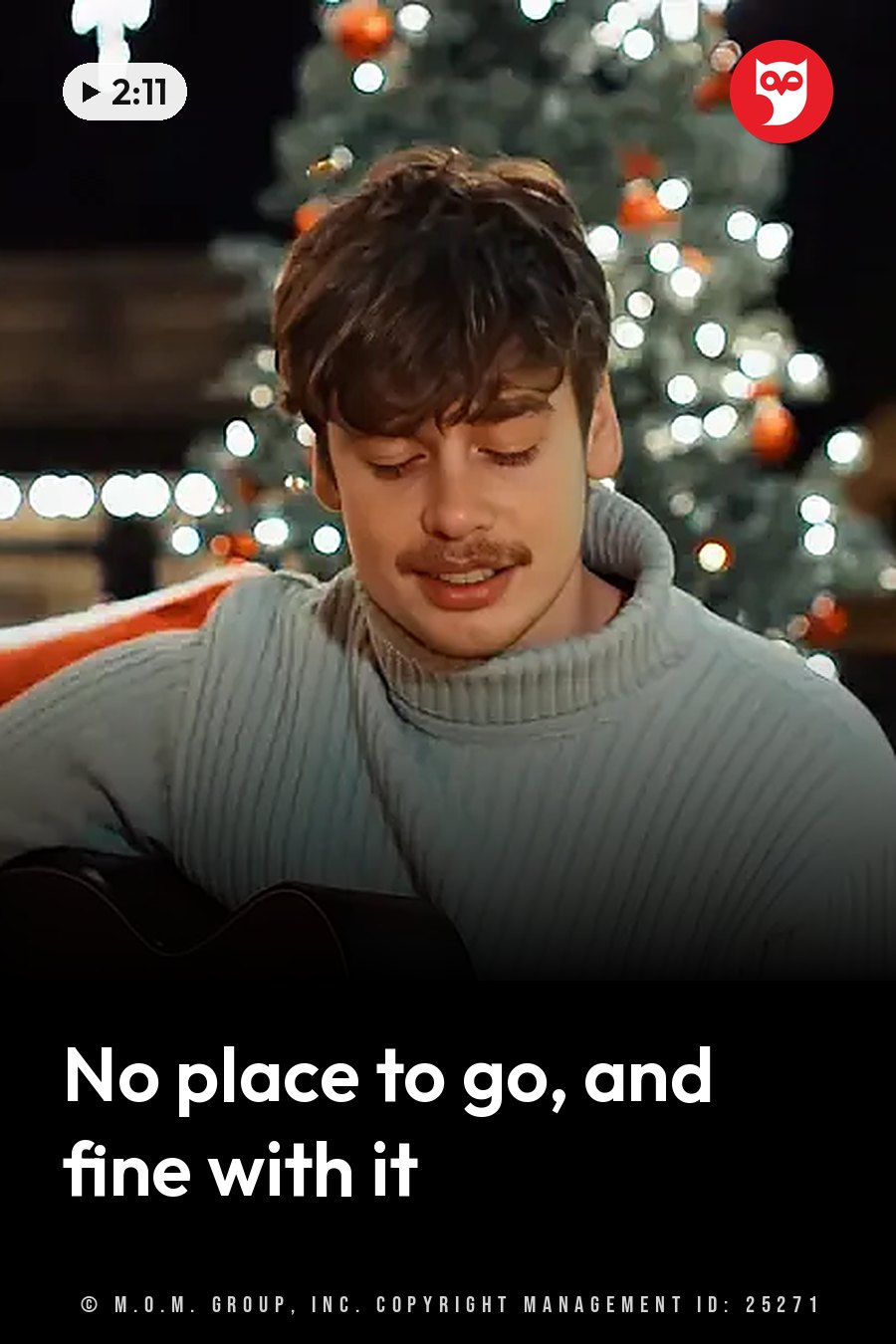 A man with a mustache in a light blue sweater stands and plays guitar in front of a lit Christmas tree at night. A headline runs in white letters across the lower third.
